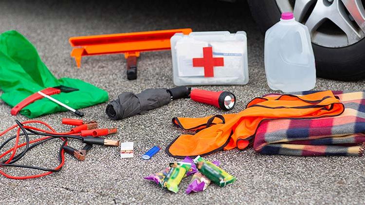 Jug of water, first aid kit, flashlight, umbrella and other emergency kit supplies upacked on the street next to a car tire.
