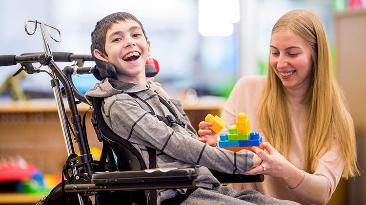 A boy in wheelchair smiles as a woman hands him toy blocks.