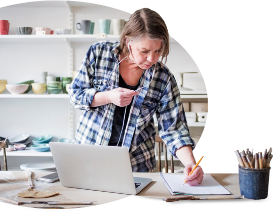 Woman in flannel shirt stands in front of a desk. She has her laptop open and is taking notes.
